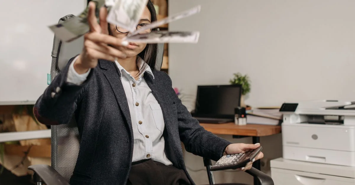 Une femme d'affaires en costard jette de l'argent tout en calculant ses finances dans un bureau moderne.