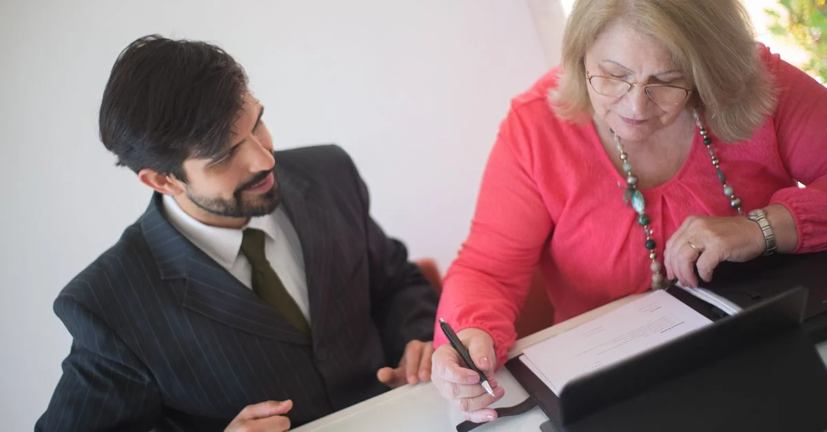 Une femme senior signant des papiers avec l'aide d'un jeune professionnel à un bureau.