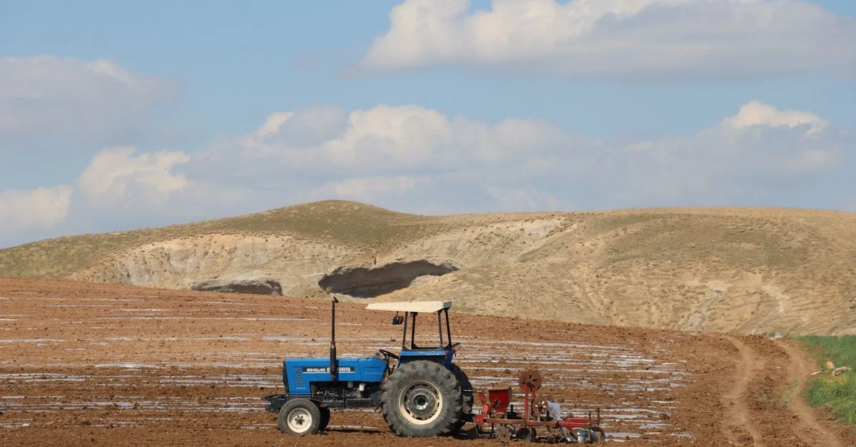 Un tracteur bleu laboure une vaste étendue de terres agricoles sur fond de collines ondulantes et de nuages.