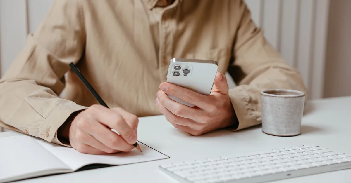 Personne prenant des notes tout en tenant un smartphone sur un bureau, illustrant le multitâche.