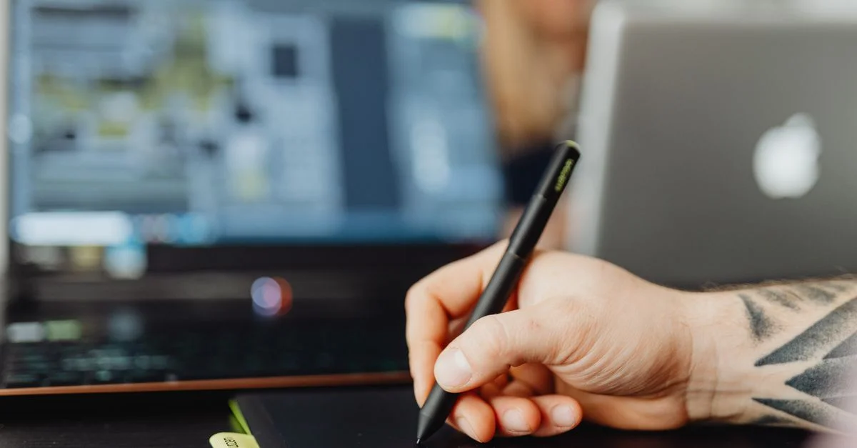 Gros plan d'une personne utilisant un stylet sur une tablette numérique avec un fond d'ordinateur portable flou.