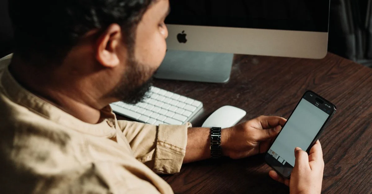 Homme travaillant sur smartphone à son bureau avec un ordinateur, illustrant la technologie dans les affaires.