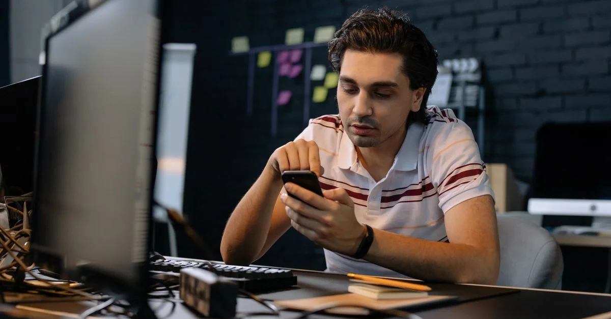 Jeune homme dans un bureau moderne utilisant un smartphone à son bureau, se concentrant sur un travail multitâche.