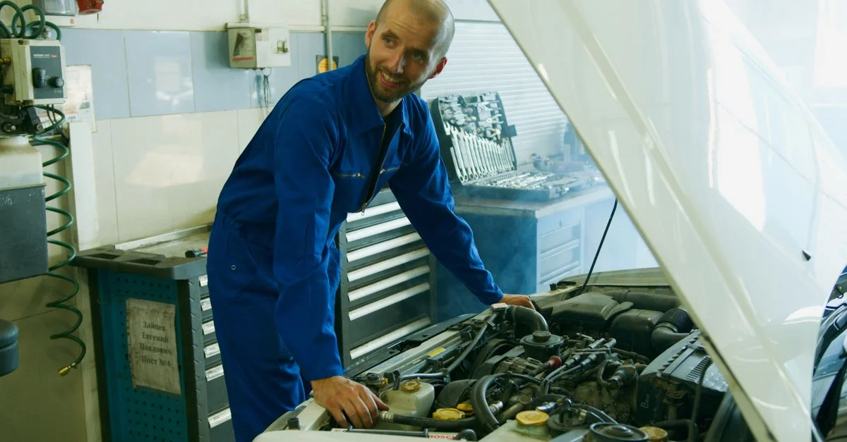 Un mécanicien souriant en salopette bleue inspectant un moteur de voiture dans un garage.
