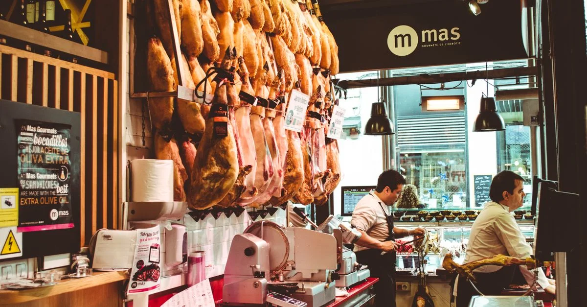 Une boutique espagnole de jambon animée avec des viandes séchées suspendues et deux hommes servant des clients.